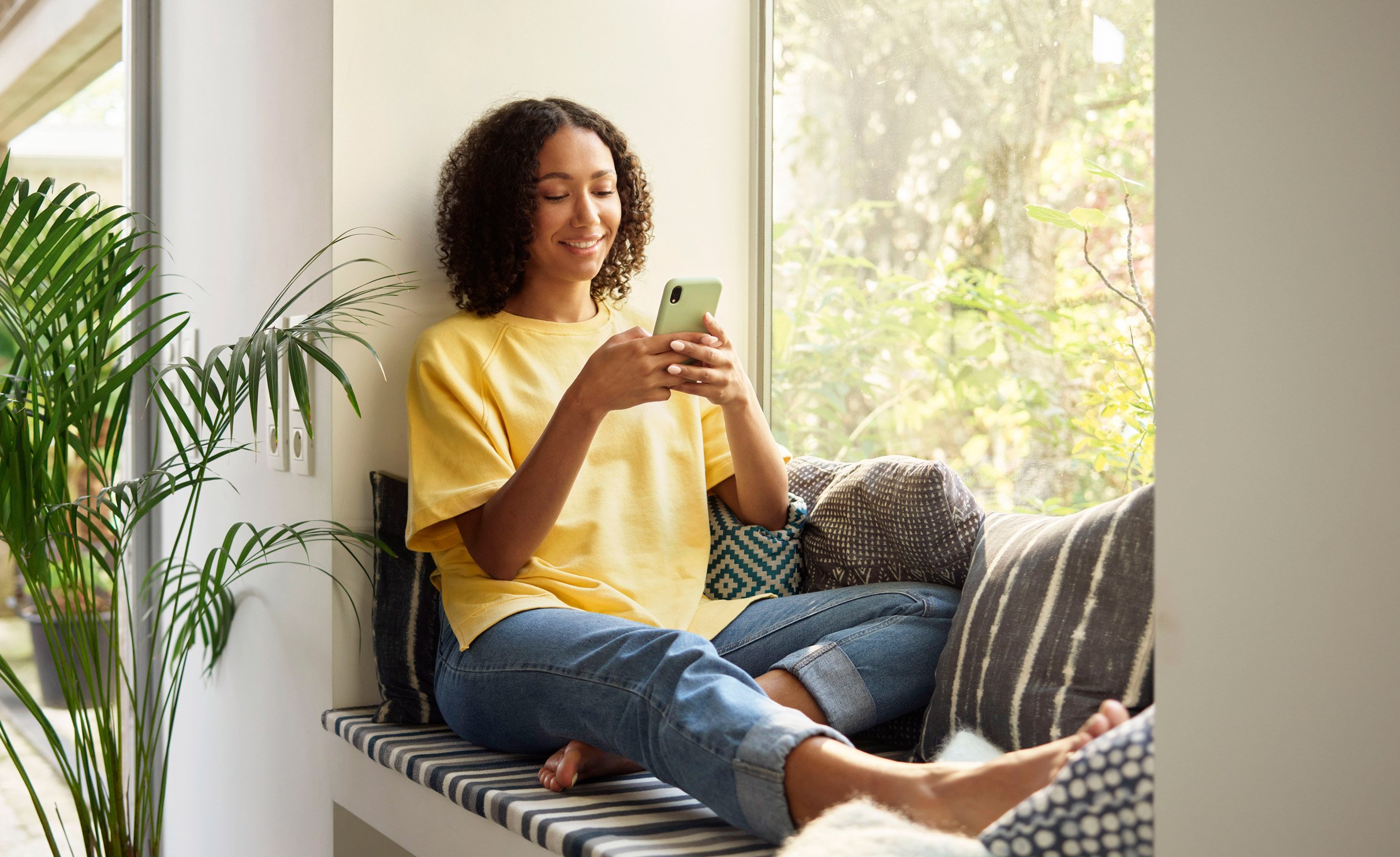 A lady sitting on her sofa by the window, holding a phone with her both hands and looking at it