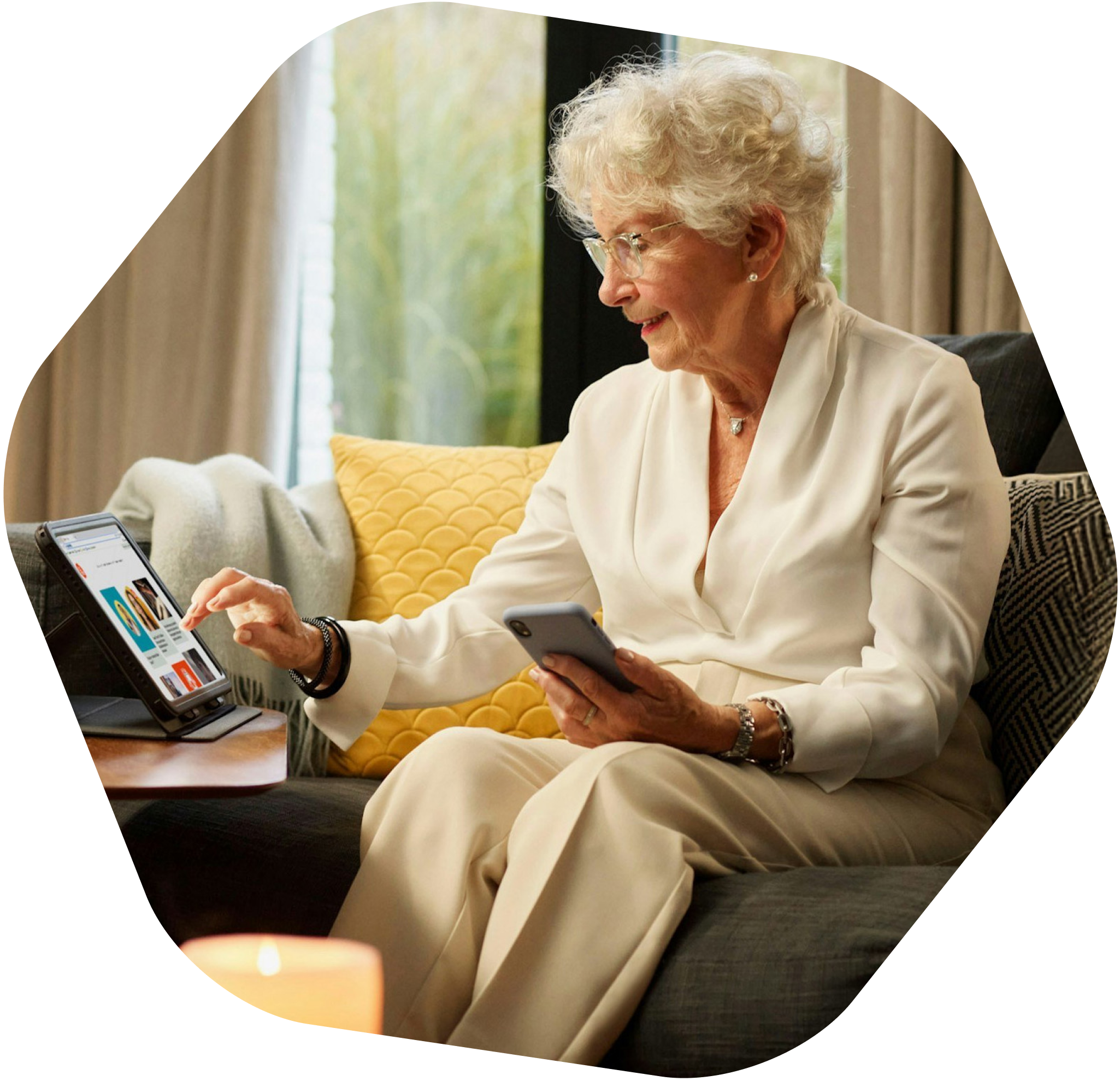 An elderly lady sitting on her sofa and using a tablet while holding her mobile phone in her left hand